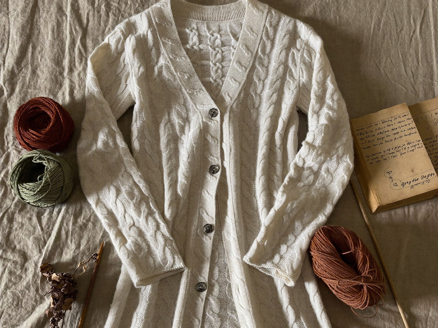 Knitting pattern flat lay — cream cable-knit cardigan with earthy-tone merino yarn, wooden needles and handwritten cable chart